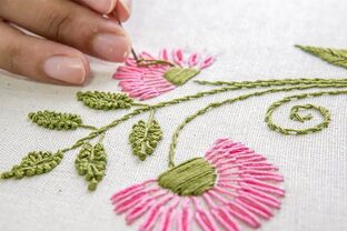 Main brodant un motif floral sur tissu blanc avec des fils verts et roses.